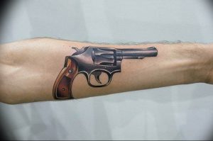 Фото тату реалистичного револьвера на мужской руке. Детализированный рисунок оружия с реалистичной передачей теней и бликов.