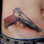 Фото тату реалистичного пистолета на мужском животе. Детализированный рисунок оружия с реалистичной передачей теней и бликов.