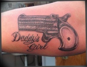Фото татуировки на руке с изображением пистолета и надписи 'Daddy's Girl'. Детализированный рисунок татуировки в черно-серых тонах.