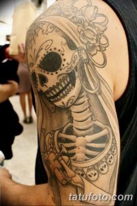 Фото татуировки на плече с изображением калаверы (черепа) в стиле Day of the Dead. Черно-белые тона с яркими цветочными акцентами, детализированная проработка, подходит для любителей мексиканской культуры.