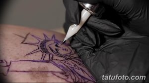 Фото процесса нанесения татуировки с изображением Статуи Свободы. Виден мастер, работающий над эскизом на коже клиента.