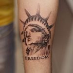 Фото тату с изображением головы Статуи Свободы и надписью "FREEDOM" на предплечье. Выполнено в черно-серых тонах, в минималистичном стиле.