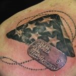 Фото тату с изображением американского флага, частично скрытого военным жетоном с гравировкой, символизирующего службу и память.