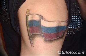 Фото тату: изображение российского флага на плече, с развевающимся полотном и реалистичными складками. Красный, синий и белый цвета.