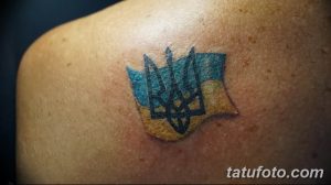 Фото тату на плече с изображением украинского герба (тризуба) на фоне флага Украины. Маленький размер, яркие цвета, патриотический символ.
