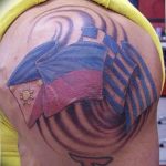 Фото тату с изображением шляпы и флага на плече, выполненное в ярких цветах с использованием синего, красного и белого. Рисунок выглядит объемно и реалистично.