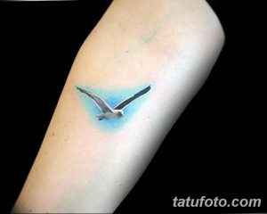 Фото акварельной татуировки на руке с изображением летящей чайки, выполненной в нежных голубых и серых тонах.