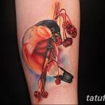 Фотография акварельной татуировки снегиря, сидящего на ветке с красными ягодами, на предплечье. Изображение выполнено в нежных, пастельных тонах.