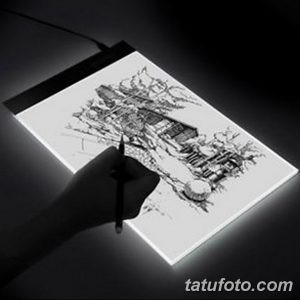 Фотография эскиза татуировки с изображением старого дома, выполненного карандашом на световом столе. Видна рука, работающая над рисунком.