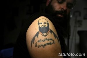 Фото тату: Портретная татуировка на плече мужчины. Изображен мужчина с бородой и традиционной одеждой, а также арабская надпись.