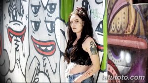 Фото тату на плече девушки: ангел с нимбом, надпись 'LOVER' и элементы граффити на фоне стены с рисунками.