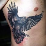 Фото тату черного ворона в полете на предплечье. Детализированный рисунок с красными брызгами краски, выполненный в реалистичном стиле.