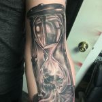 Черно-серое фото татуировки песочных часов, в которых вместо песка изображен череп, на руке.