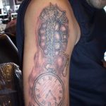 Фото тату на плече, изображающее карманные часы, пачку денег и надпись 'Time is Money'. Реалистичный рисунок в черно-серых тонах.