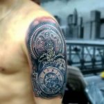 Фото татуировки на плече, изображающей механические часы с видимыми шестеренками и деталями. Татуировка выполнена в черно-серых тонах.
