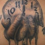Фото тату на теле, изображающее руку, словно выходящую из циферблата часов, с каплями, стекающими вниз, выполненное в темных тонах.