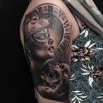 Фото тату волка и часов, выполненное в черно-серых тонах на плече. Реалистичный рисунок с детальной проработкой шерсти волка и механизма часов.