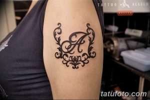 Фото тату с элегантной монограммой, состоящей из буквы 'А', обрамленной декоративными элементами. Черно-белый рисунок на плече.