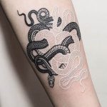 Фото тату: две змеи, одна черная, другая белая, обвиваются вокруг руки, образуя сложный узор. Детализированная проработка чешуи и глаз.
