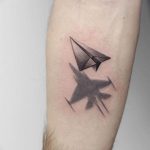 Черно-серое фото татуировки на предплечье с изображением бумажного самолетика и военного истребителя, символизирующего мечты и реальность.