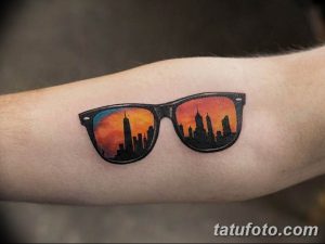 Фото тату в виде очков, в линзах которых отражается силуэт города на фоне заката. Яркая, красочная татуировка на руке, выполненная в стиле акварель.