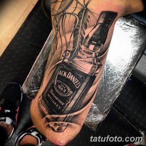 Фото тату на руке с реалистичным изображением бутылки виски Jack Daniel's. Черно-серый рисунок с высокой детализацией.