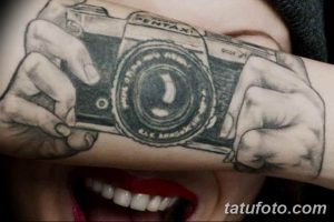 Черно-белое фото татуировки с изображением старинного фотоаппарата Pentax, держащегося в руках. Детализированный рисунок на предплечье.