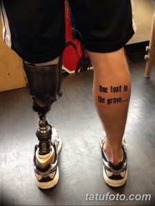 Фото тату на голени с надписью 'One foot in the grave...', выполненной черными чернилами. Рядом с татуировкой - протез ноги.