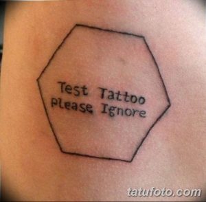 Фото тату на руке: черно-белая надпись 'Test Tattoo please Ignore' в шестиугольнике. Простой, минималистичный рисунок татуировки.