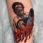 Фото татуировки на ноге с изображением Leatherface из фильма 'Техасская резня бензопилой', держащего бензопилу. Рисунок выполнен в ярких цветах.