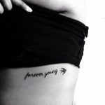 Фото тату надписи 'forever young' на боку. Черный контур, тонкий шрифт, минималистичный дизайн. Идеально для тех, кто ценит простоту и символизм.