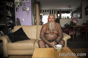 Фотография пожилого мужчины с татуировками на теле, сидящего на диване в домашней обстановке, демонстрирующая его индивидуальность и любовь к искусству.