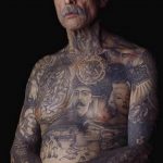 Фотография пожилого мужчины с множеством татуировок на теле, демонстрирующих его богатый жизненный опыт.