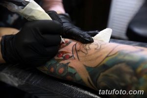 Фото тату: мастер тату наносит татуировку на руку клиента. Виден тату-аппарат и рука клиента с уже нанесенным рисунком.