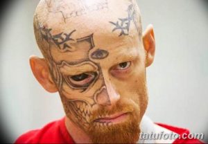 Фото татуировки на лице мужчины: зловещая маска с острыми зубами и пронзительным взглядом, выполненная в черно-белой гамме. Дополнительные татуировки на лбу.