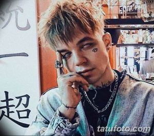 Фото тату: стилизованный глаз с паутиной под ним, выполненный в черных тонах на лице молодого человека. Детализированный рисунок татуировки.