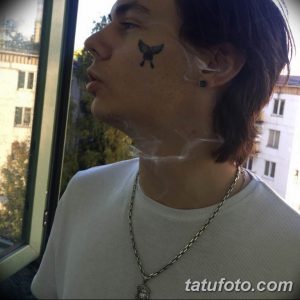 Фотография мужчины с небольшой татуировкой птицы под глазом. Демонстрация минималистичного стиля и изящного дизайна.