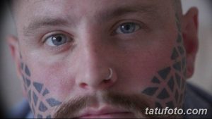 Фотография мужчины с геометрическими татуировками, покрывающими его щеки и часть лба. Татуировки выполнены в сером цвете и имеют сложный узор.