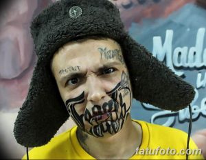 Фото тату: яркая татуировка на лице мужчины в стиле олдскул с изображением черепа и надписями. Необычный и смелый выбор для самовыражения.