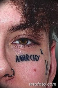 Фото тату: надпись 'ANARCHY' и другие символы, выполненные черными чернилами на лице мужчины. Провокационный дизайн и бунтарский дух.