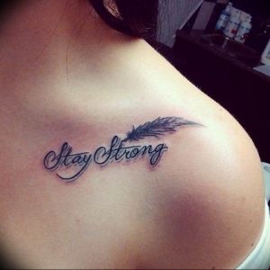 Фото тату на ключице девушки. Изображение надписи 'Stay Strong' с изображением пера. Татуировка выполнена в черном цвете.