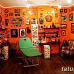 Фото тату-студии с оранжевыми стенами, усыпанными эскизами татуировок. Зеленое кушетка и множество инструментов для тату.