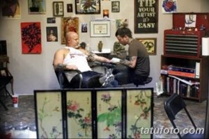 Фото процесса нанесения татуировки: мастер работает над рисунком на руке клиента. В студии много картин и декоративных элементов.