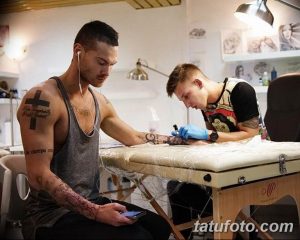 Фото процесса нанесения татуировки: мастер в черной одежде работает над рисунком на руке клиента, клиент смотрит в телефон.