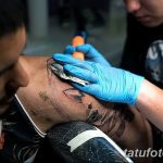 Фото процесса нанесения татуировки: мастер в голубых перчатках работает над рисунком на плече клиента, используя тату-машинку.