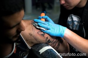 Фото процесса нанесения татуировки: мастер в голубых перчатках работает над рисунком на плече клиента, используя тату-машинку.