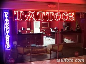 Фото тату салона с яркой неоновой вывеской 'TATTOOS'. Внутри видно мебель для ожидания и ресепшн. Современный дизайн.