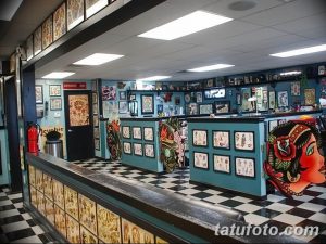 Фото тату салона с черно-белой плиткой на полу. Видны стойка ресепшн, кресла для ожидания и стены, украшенные эскизами татуировок.