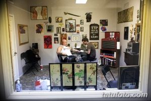 Фото тату процесса нанесения рисунка. Мастер работает над татуировкой на руке клиента. В студии много картин и декоративных элементов.