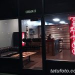 Фото тату салона ночью. Яркая неоновая вывеска 'TATTOO' привлекает внимание. Внутри видно интерьер и посетителей. Современный дизайн.